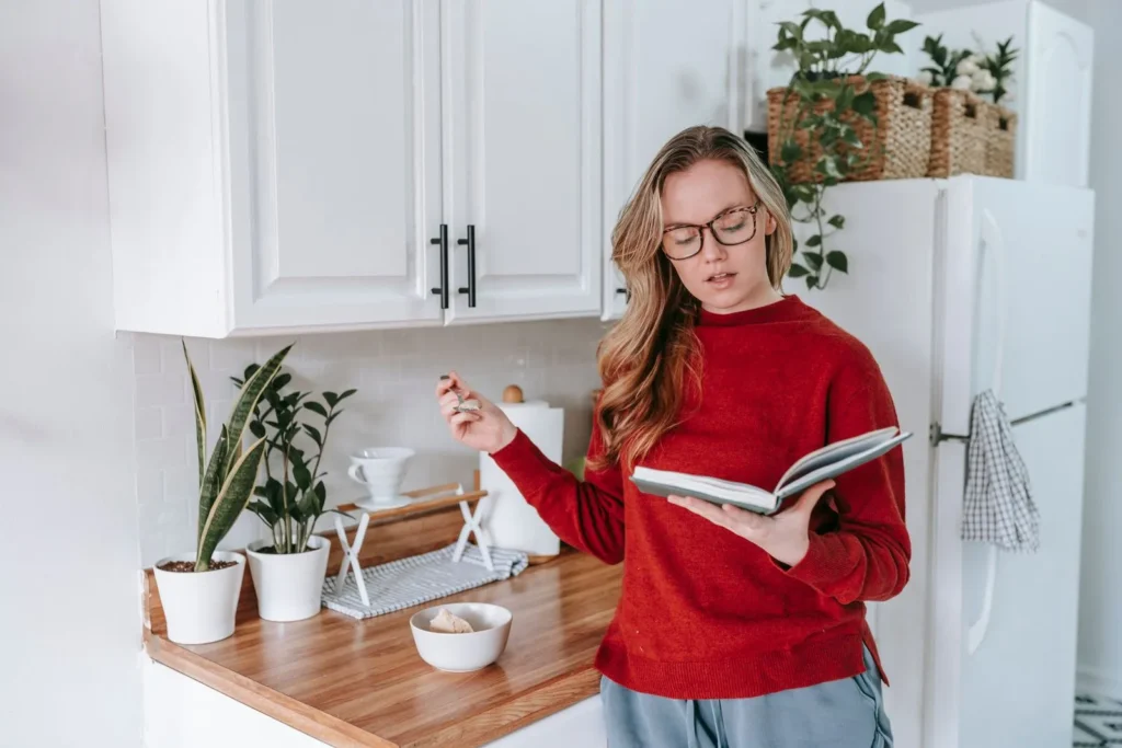 Woman in glasses reading a book in a bright kitchen with potted plants